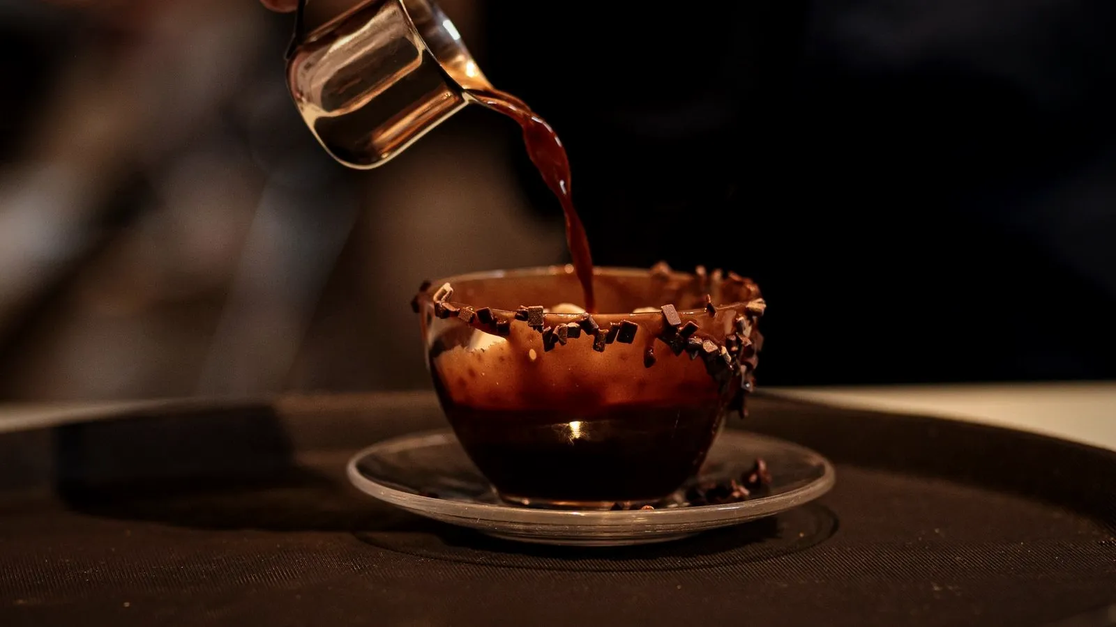 Coffee being poured from a small metal pitcher into a glass cup rimmed with chocolate pieces