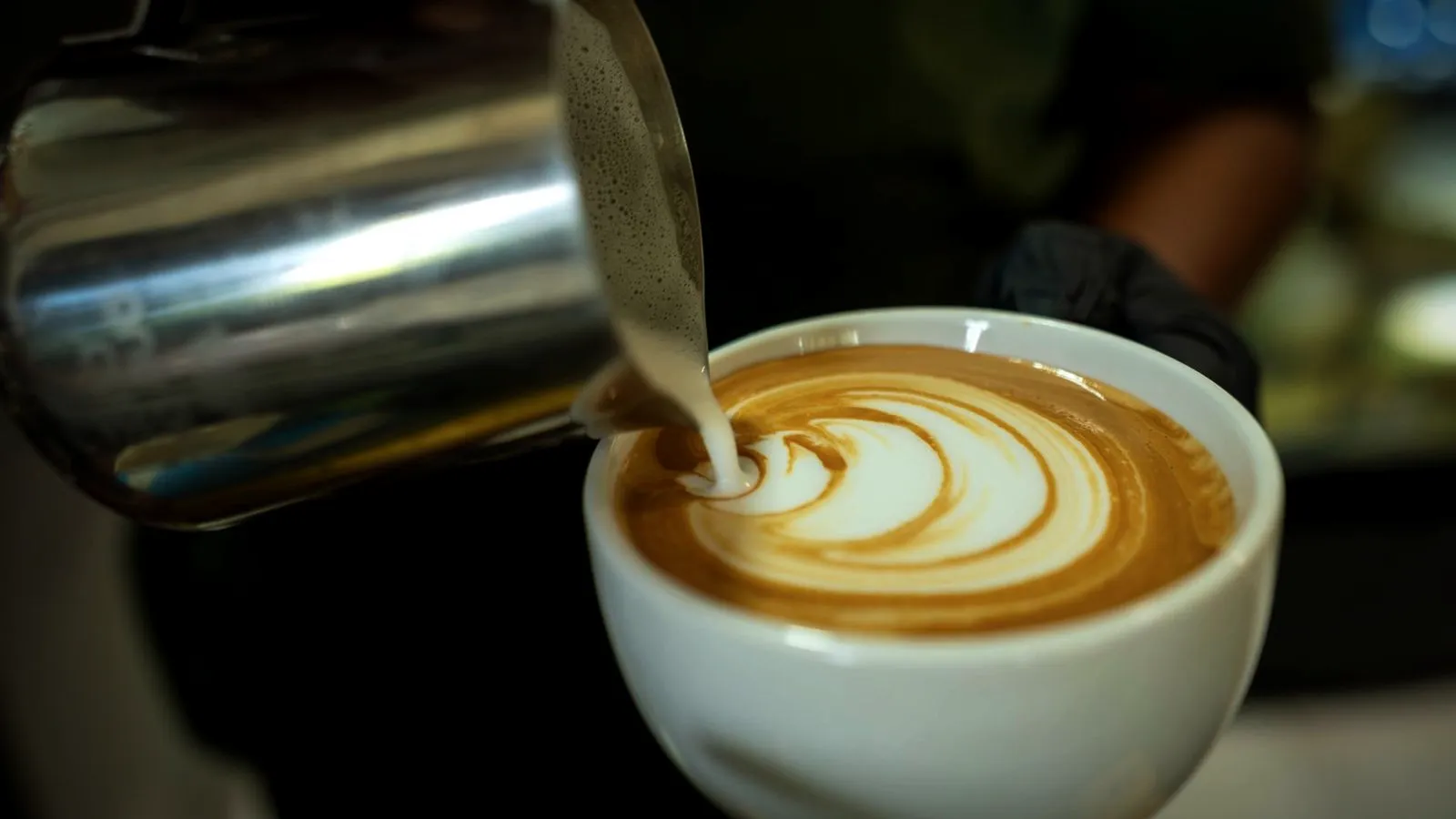Silky steamed milk being poured from a stainless pitcher into espresso, forming latte art in a white cup