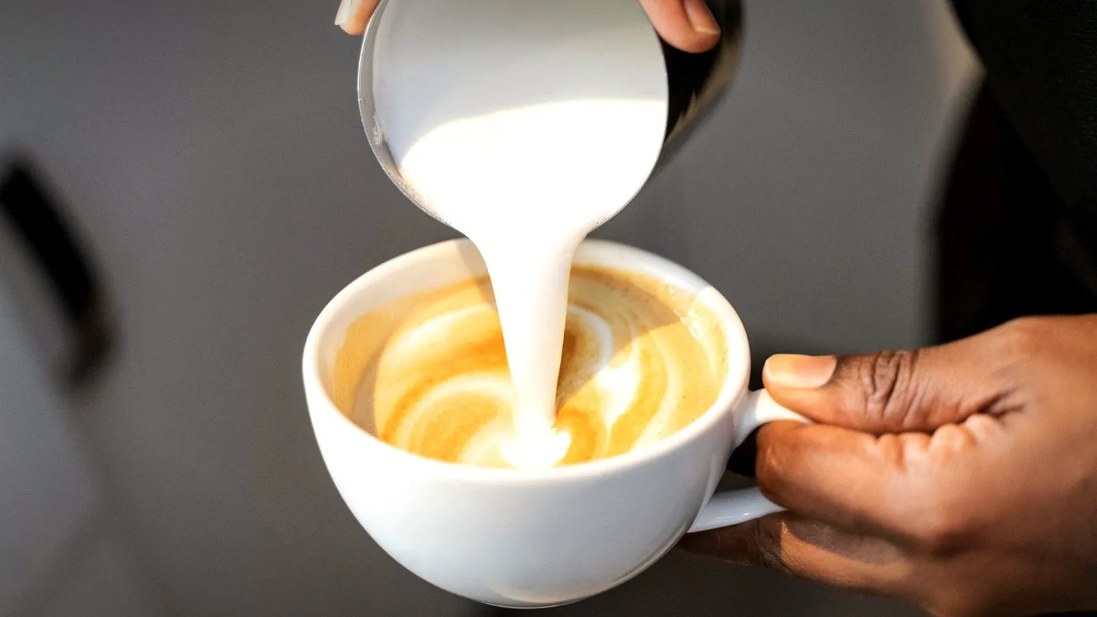 A thick stream of steamed milk poured from a pitcher into a white cup of espresso