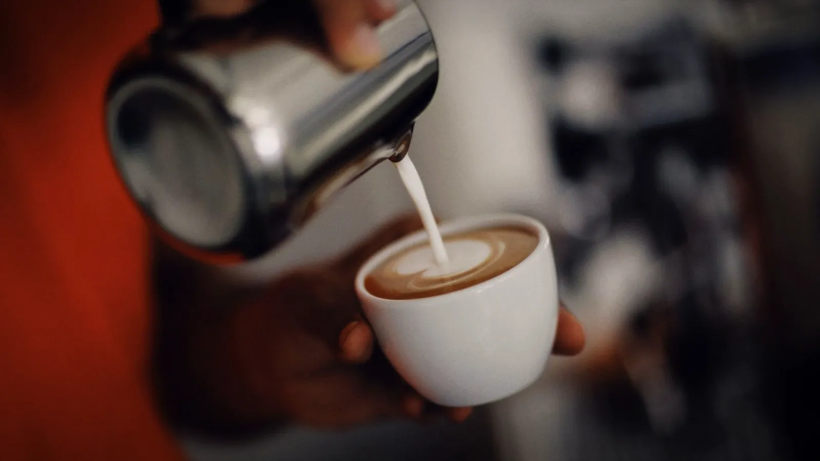 Pouring steamed milk from a steel pitcher into a cup of espresso to form a foam pattern