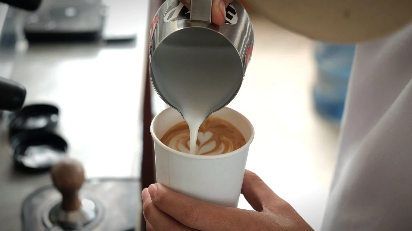 A barista pouring steamed milk from a stainless pitcher into a white paper cup, forming latte art