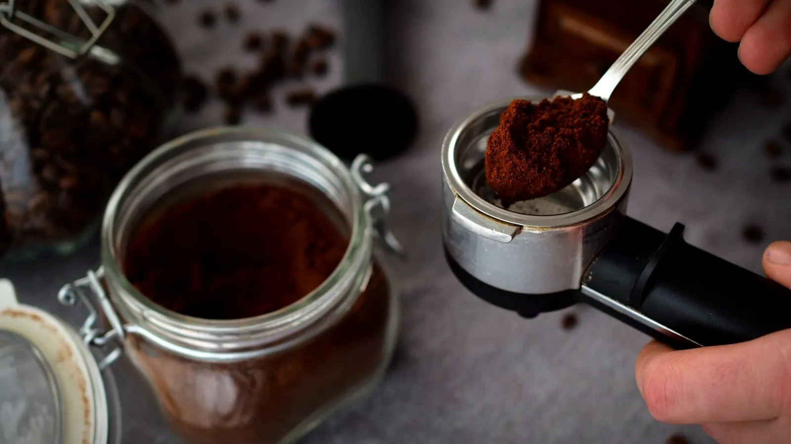 Pre-ground coffee being spooned from a glass jar into an espresso portafilter