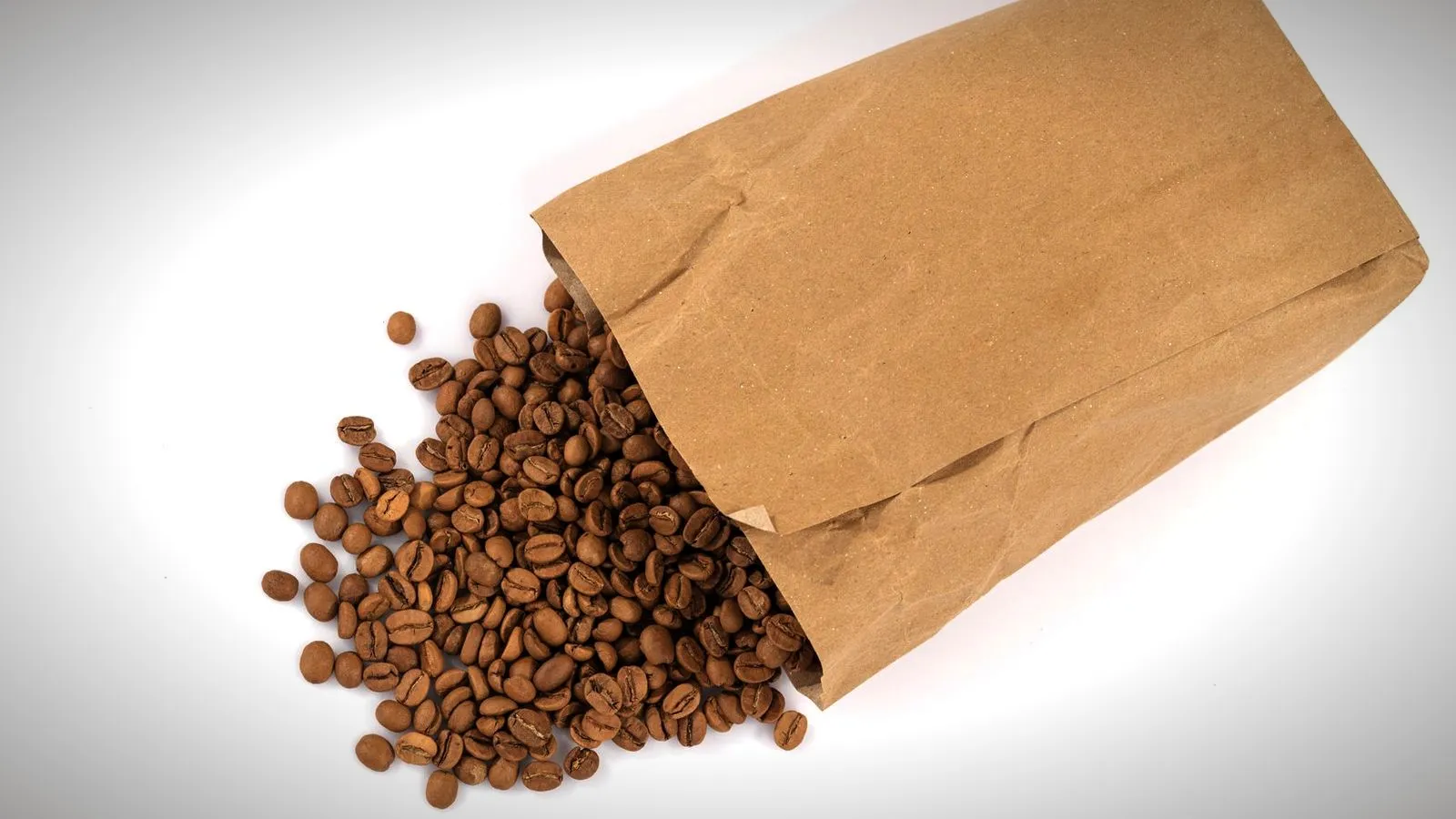 Roasted coffee beans spilling from a plain kraft paper bag onto a white surface