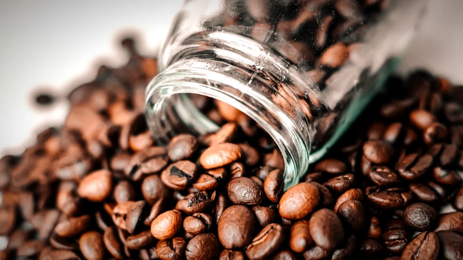 Roasted coffee beans spilling from a glass jar onto a light surface