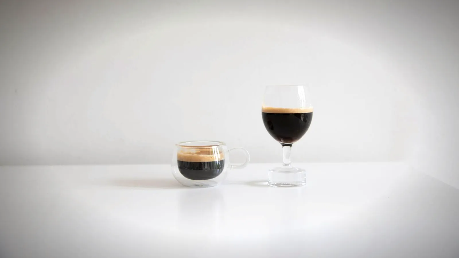 A short espresso in a small glass cup next to a taller stemmed glass of black coffee