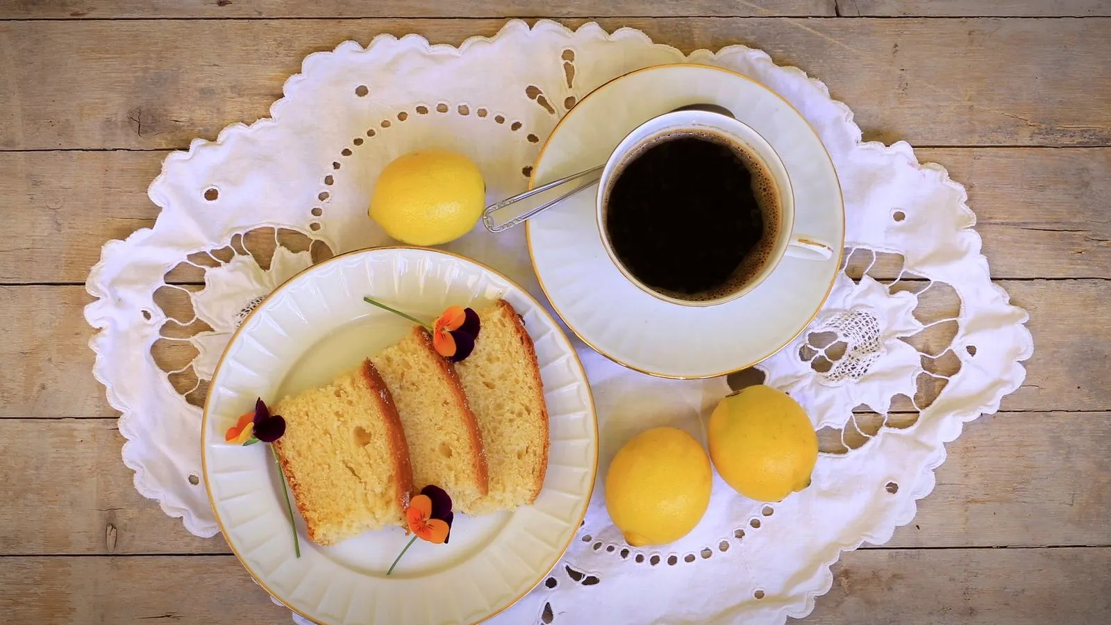 Top view of a small cup of black coffee on a lace doily with whole lemons and a plate of lemon cake