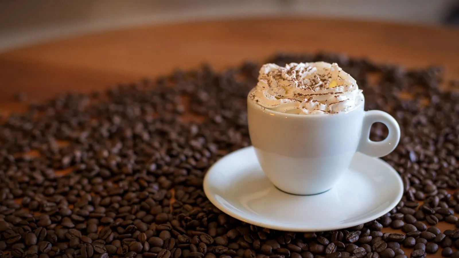 Small white cup topped with whipped cream and chocolate shavings on a saucer, surrounded by roasted coffee beans