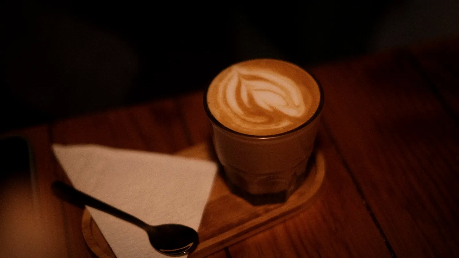A small glass of milk coffee with latte art on a wooden tray beside a napkin and spoon