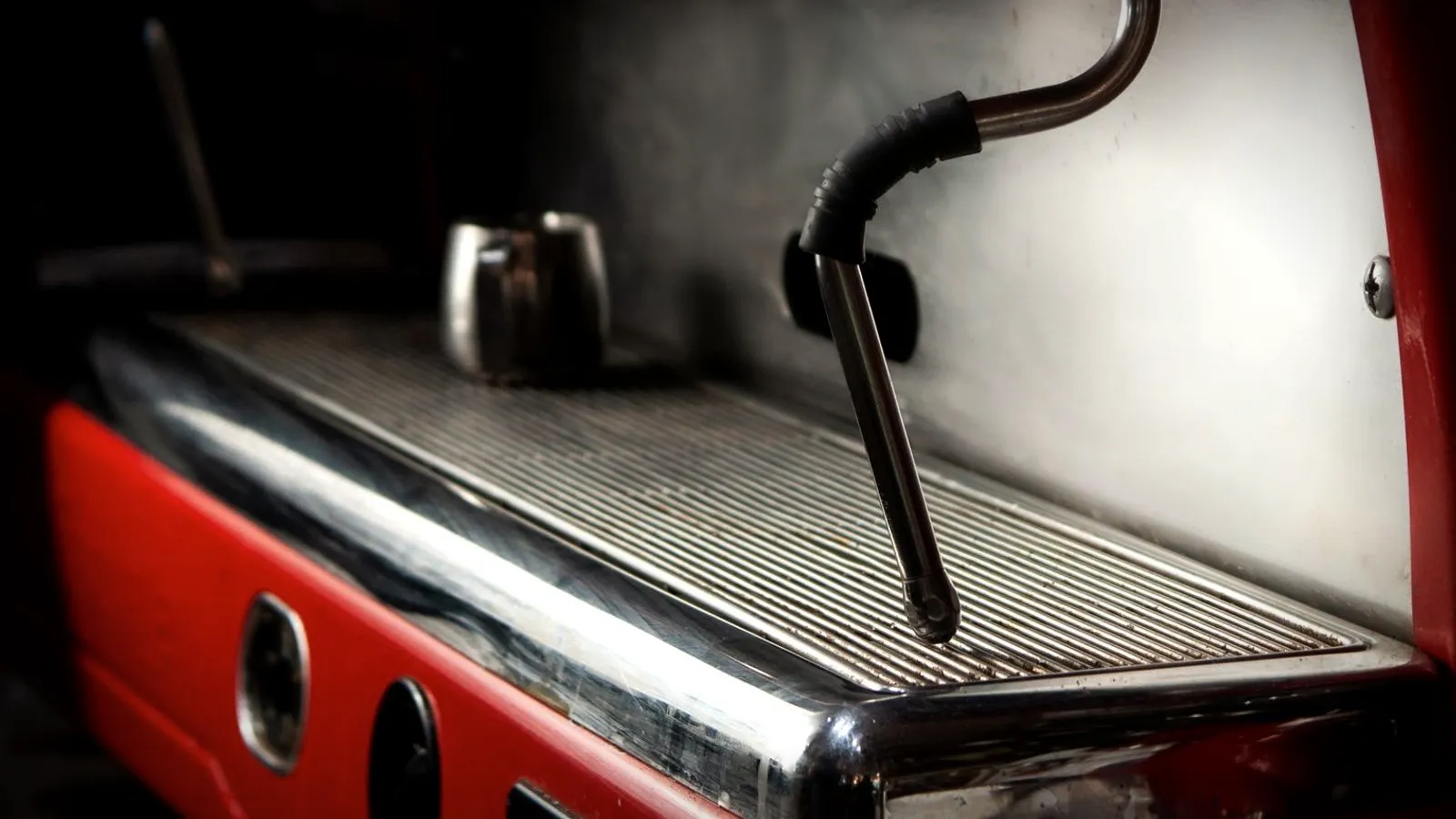 The steam wand and drip tray of a red espresso machine, with a small milk pitcher at the back