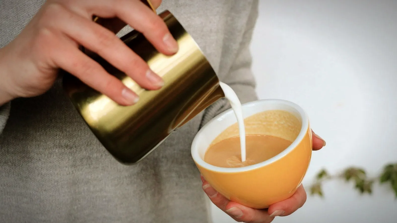 Steamed milk poured from a gold-colored pitcher into a yellow cup of coffee