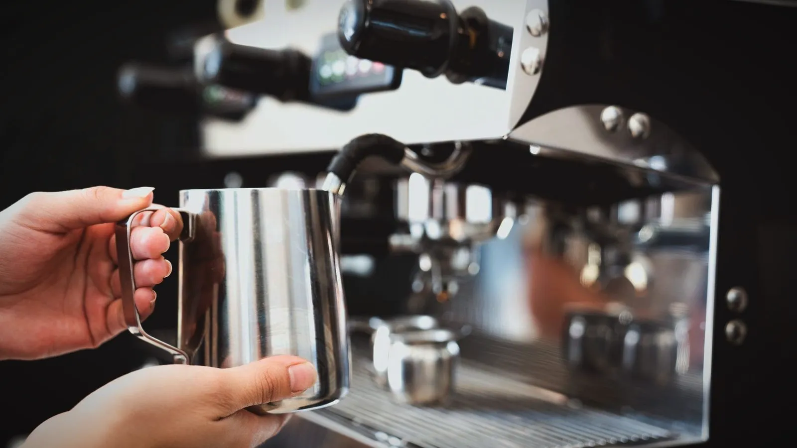 Hands holding a stainless steel milk pitcher under the steam wand of a commercial espresso machine