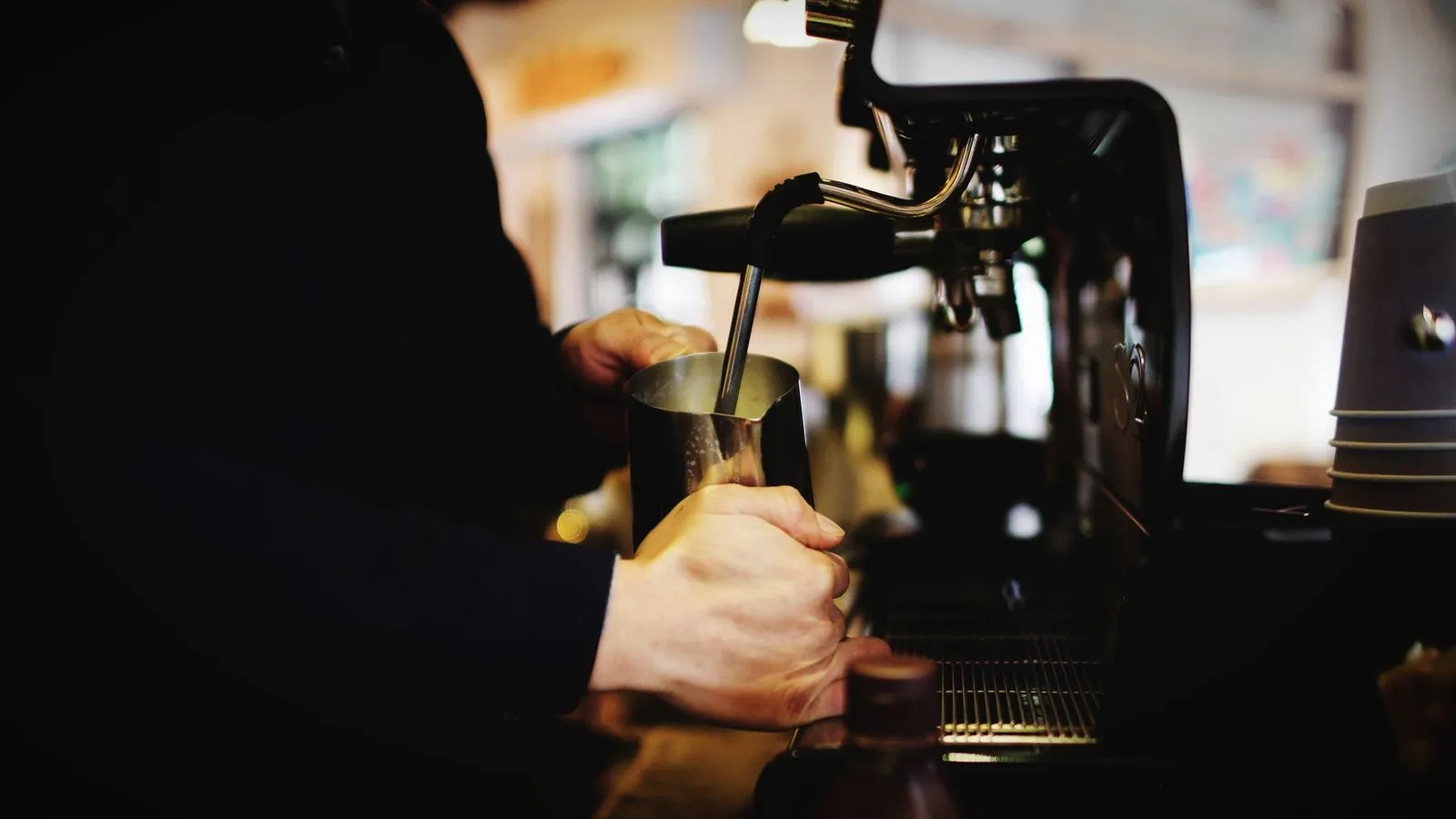 Barista holding a steel pitcher under the steam wand to steam milk, in a dim cafe