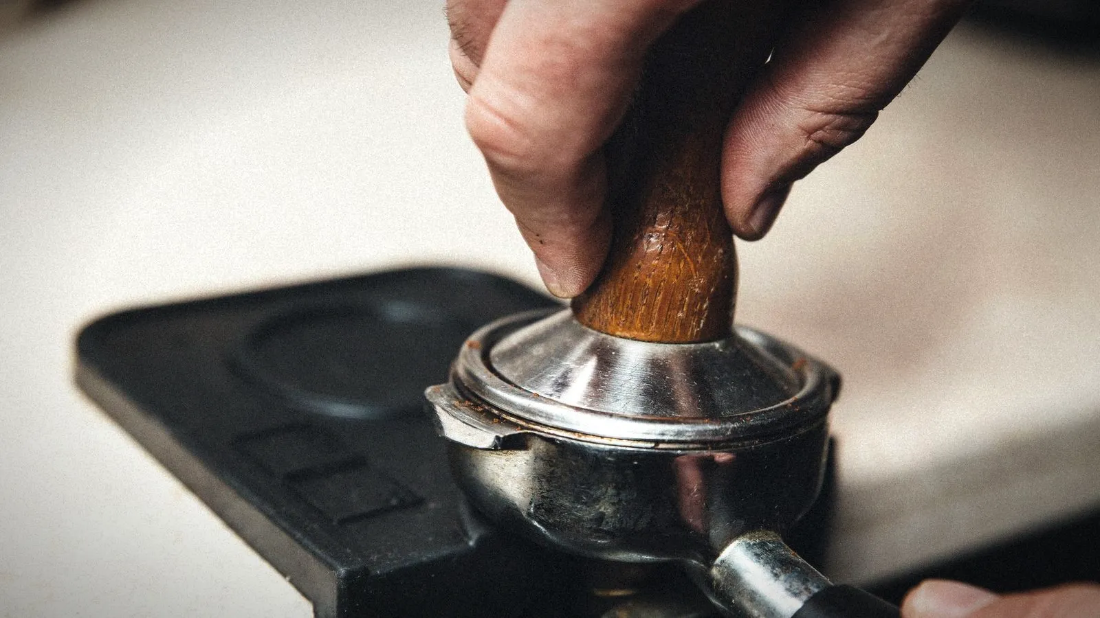 Hand pressing a wooden-handled tamper into coffee in a portafilter on a tamping mat