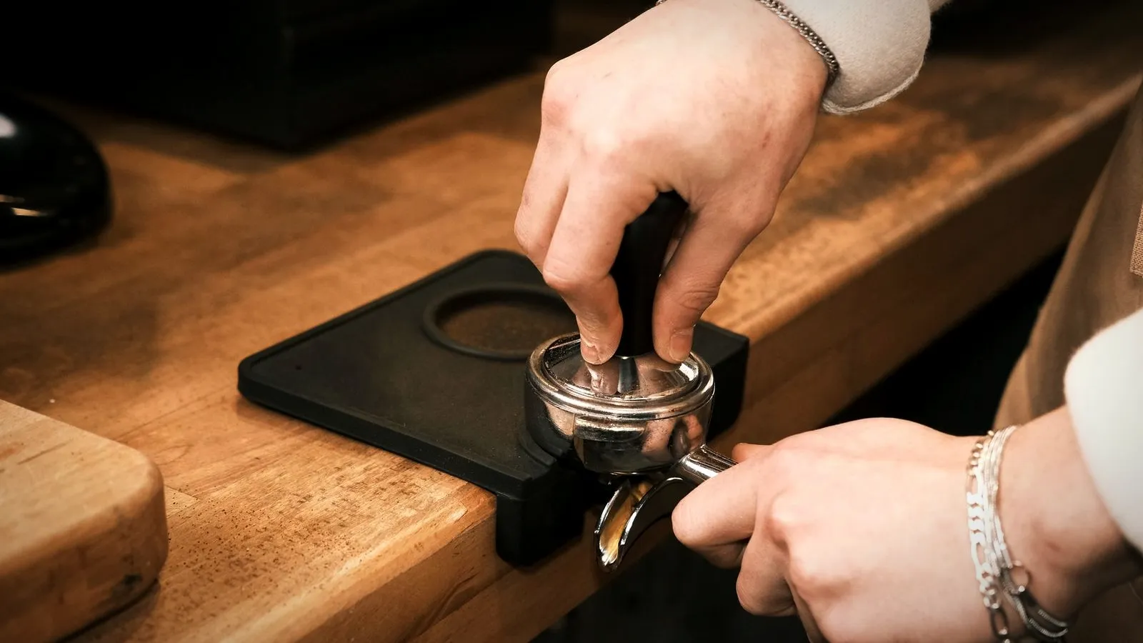 Hands tamping coffee in a portafilter that rests on a black rubber tamping mat on a wooden counter
