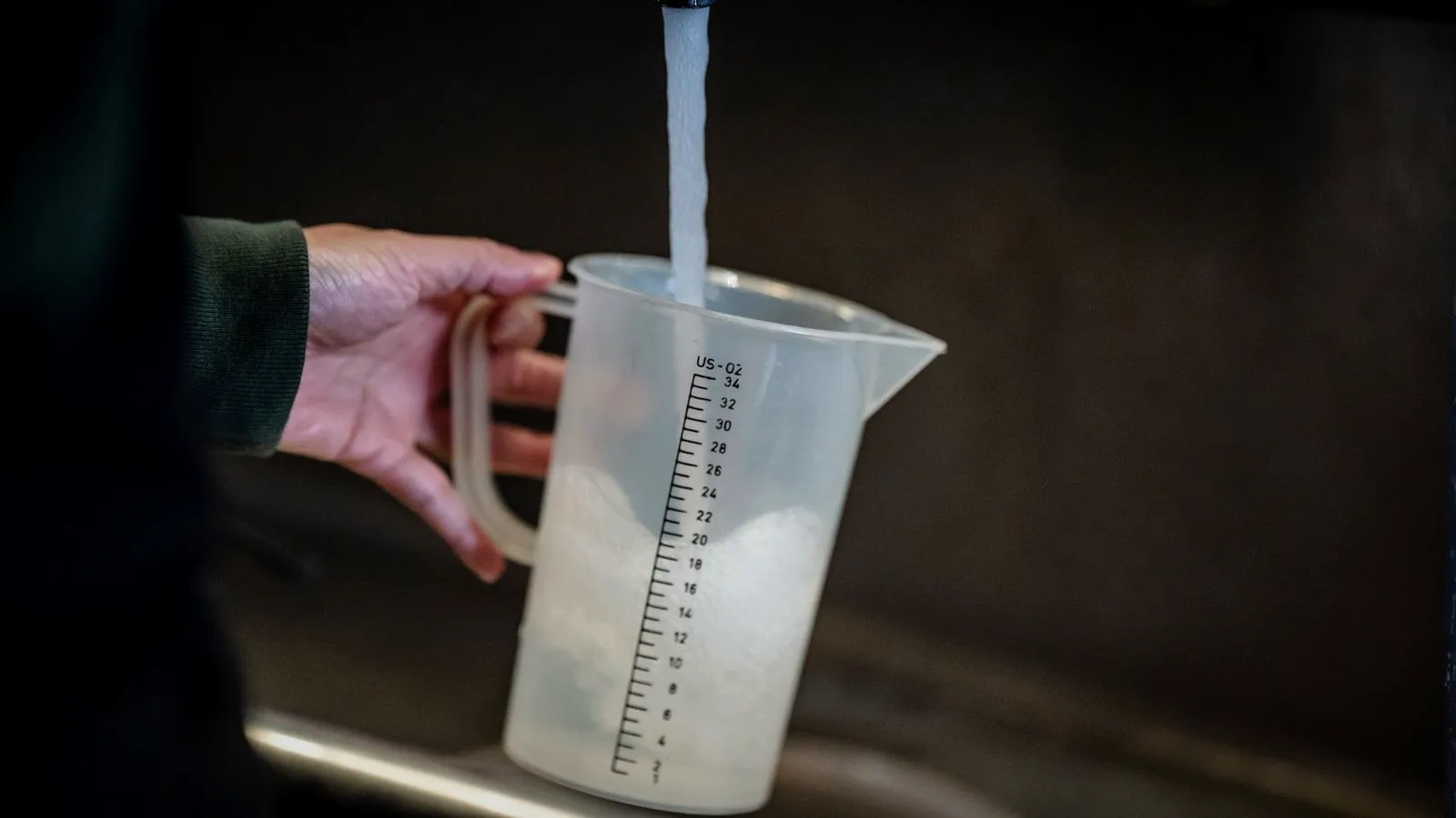 Hand filling a plastic measuring jug with tap water at a kitchen faucet