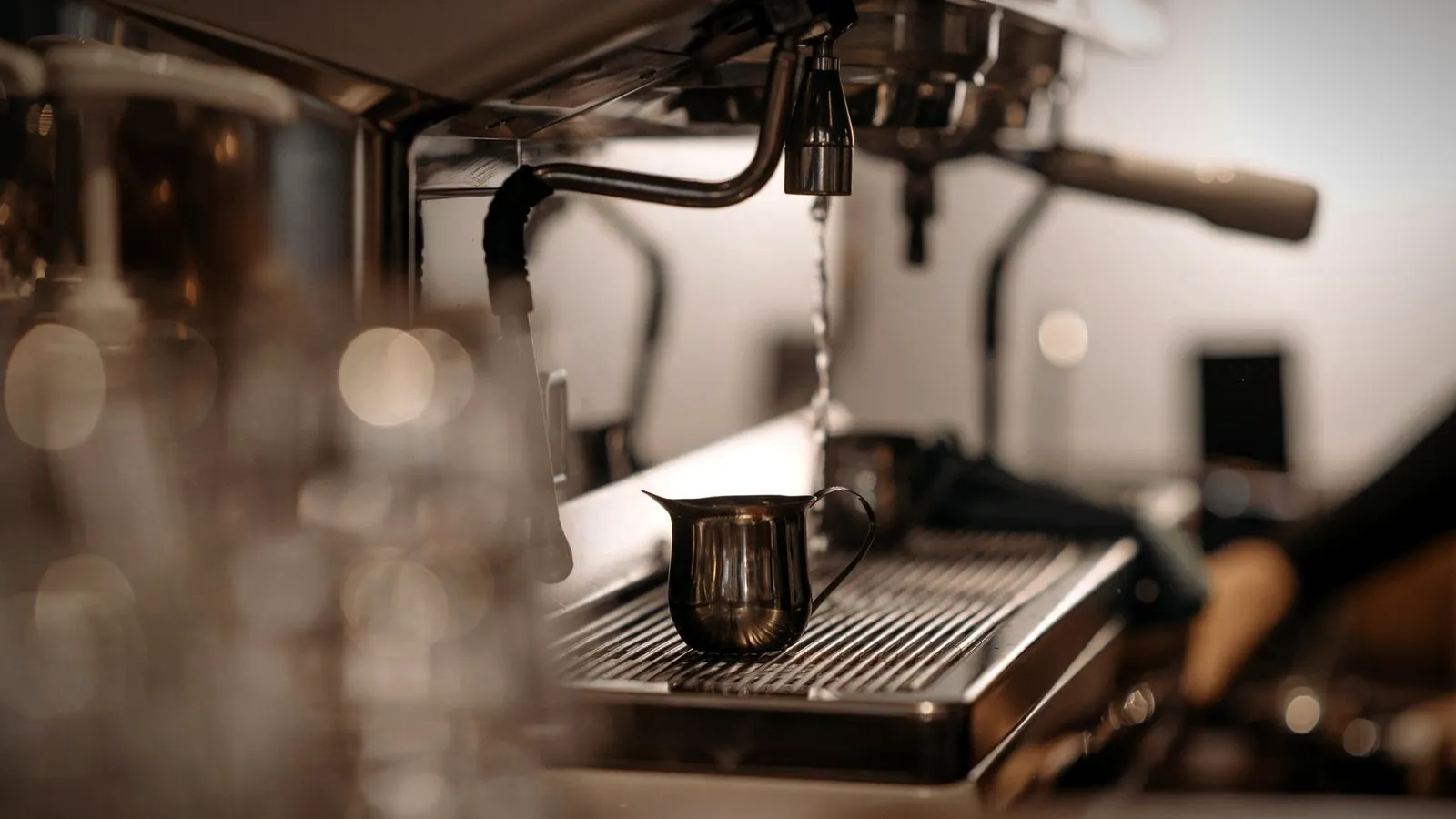 Thin stream of hot water running from a cafe espresso machine onto the drip tray, a small steel pitcher beside it