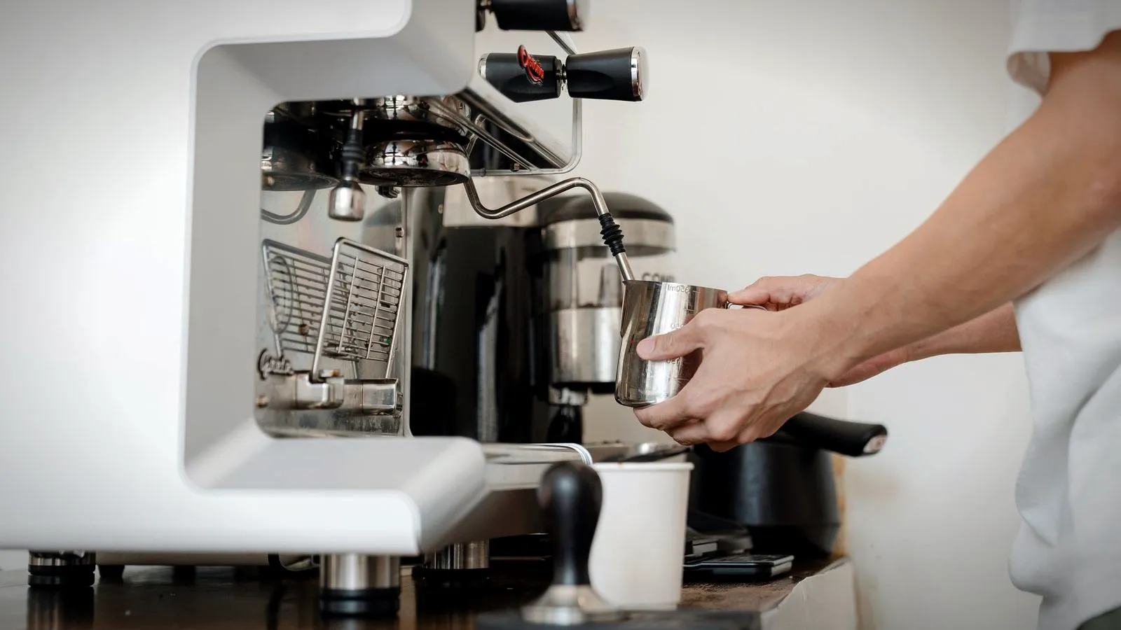 Side view of a white espresso machine with a hand steaming milk in a steel pitcher under the steam wand
